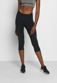 Schwarze Capri-Leggings aus dehnbarem Stoff, mit strukturierten Seiteneinsätzen, einem hohen Taillenbund und kombiniert mit schwarzen Sportschuhen.