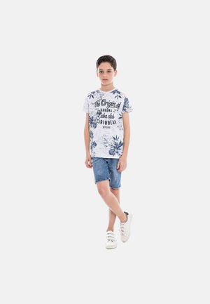 Garçon debout, jambes croisées, portant un t-shirt blanc avec un imprimé graphique tropical bleu, un short en denim et des baskets blanches sur fond uni.