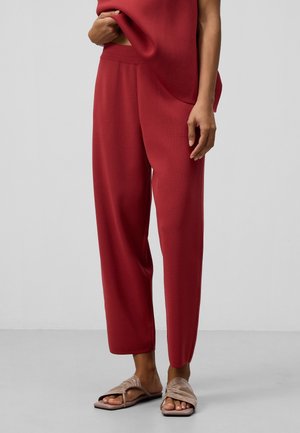 Osoba ubrana w luźne czerwone spodnie i beżowe płaskie sandały z odkrytymi palcami, stojąca na tle jednolitego jasnoszarego tła.