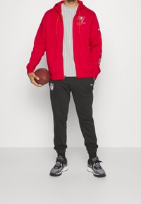 Rød zip-up hoodie med logo, grå stribet skjorte, sorte sweatpants med NFL-logo og sorte løbesko, mens han holder en fodbold.