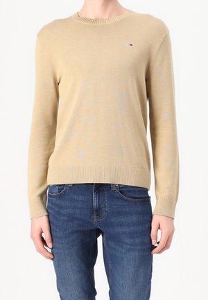 Pull beige à manches longues avec col rond et petit logo Tommy Hilfiger sur la poitrine, porté avec un jean denim bleu sur une personne debout.