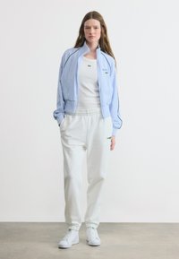 Νεαρή γυναίκα στέκεται με τα χέρια στις τσέπες, φορώντας ανοιχτό μπλε ζακέτα με φερμουάρ, λευκή φανέλα, λευκή φόρμα και λευκά αθλητικά παπούτσια, μπροστά σε μονόχρωμο φόντο.