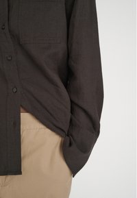 Dark brown button-up shirt with a front pocket, made of a textured fabric, paired with khaki pants. The shirt has a relaxed fit and long sleeves.