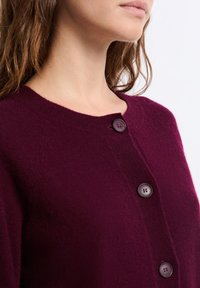 Frau in einer weichen, tiefvioletten Knopfkarte, die den Ausschnitt und den oberen Brustbereich an einem schlichten hellen Hintergrund zeigt.