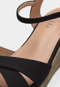 Sandale compensée noire avec brides croisées en tissu et bride à la cheville sur une semelle intérieure beige et un talon plateforme tissé.