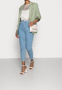 Blazer verde claro, top de satén beige, jeans azul claro y sandalias blancas con tacón cuadrado. Pequeña bolsa de hombro beige con acentos dorados.