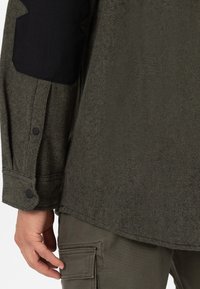 Camicia verde oliva con tessuto texturizzato, toppe nere sulle spalle, polsini con bottoni e orlo dritto. Abbinata a pantaloni stile cargo.
