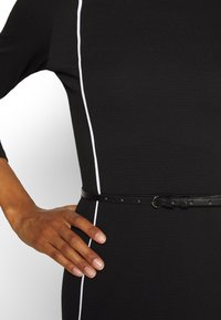 Robe noire avec des surpiqûres blanches sur les côtés, complétée par une ceinture en cuir noir avec des clous en métal. Met l'accent sur une coupe ajustée et une texture lisse.