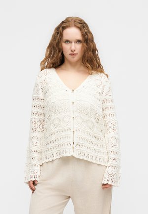 Junge Frau mit langen lockigen Haaren, die eine weiße, gehäkelte Strickjacke mit Perlenknöpfen und beige Hosen trägt, steht vor einem einfarbigen Hintergrund.