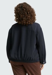 Veste bleu marine en tissu lisse, dotée d'un ourlet élastique froncé, d'épaules basses et de poignets à boutons-pression. Vue arrière montrée.