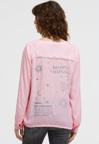 Frau mit lockigem Haar, die ein langärmliges Pinkes Shirt mit inspirierendem Text und himmlischen Illustrationen auf dem Rücken trägt, kombiniert mit dunkler Jeans.