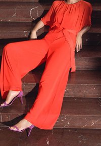 Röd byxdress med vida ben, samlad halsringning och knytbar midja, i kombination med lila högklackade sandaler på marmorgtrappor.