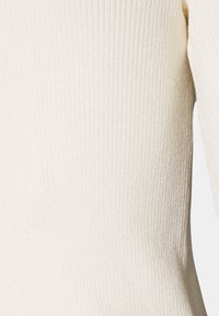 Top a righe bianche in maglia con maniche lunghe. Il tessuto ha un aspetto morbido e testurizzato, con una leggera lucentezza e una silhouette aderente.