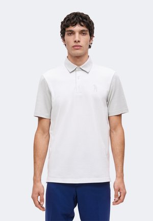 Homme aux cheveux bouclés foncés portant un polo gris clair à manches courtes et un pantalon bleu marine, debout sur un fond uni.