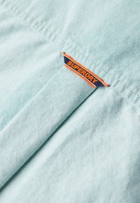 Tessuto azzurro chiaro con una trama testurizzata, con un'etichetta "SUPERDRY" arancione e blu ben visibile. I punti di cucitura sono ordinati e paralleli.