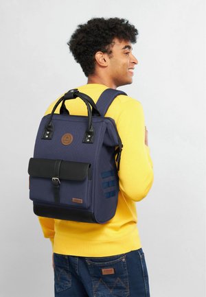 Marineblaue Tasche mit schwarzen Akzenten, strukturiertem Stoff, vorderer Klappe und Schulterriemen. Entworfen mit einem Logo und Riemenverzierungen an der Seite.