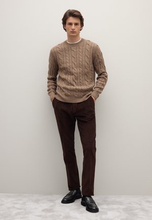 Pull en maille torsadée couleur marron clair, associé à un pantalon en velours côtelé marron foncé et des mocassins noirs. Le modèle se tient avec les mains dans les poches.
