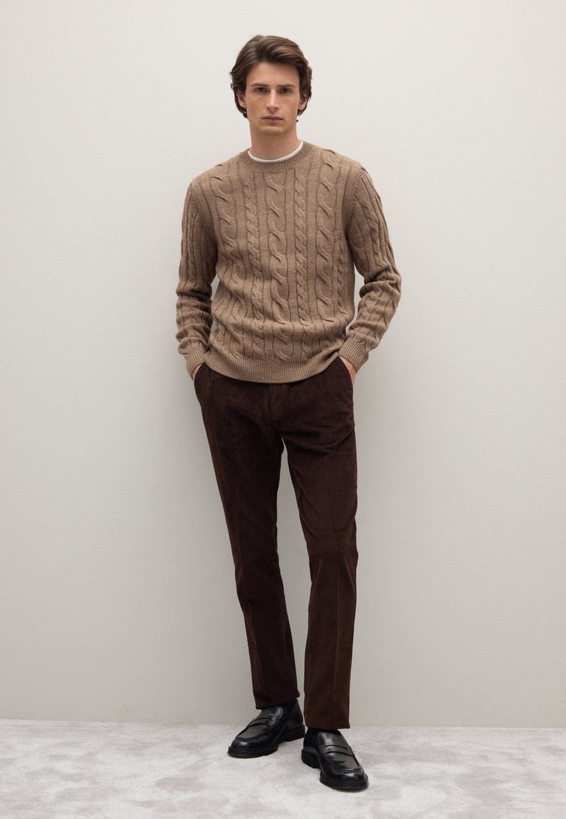 Hellbrauner Strickpullover, kombiniert mit dunkelbraunen Cordhosen und schwarzen Slippern. Das Model steht mit Händen in den Taschen.