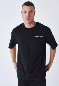 Zwart katoenen t-shirt met korte mouwen, voorzien van witte tekst "PROJECT X PARIS" op de linkerkant van de borst. Relaxte pasvorm, gladde textuur.