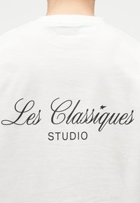 Οπίσθια όψη ατόμου που φοράει λευκό πουκάμισο με μαύρο κείμενο "Les Classiques" σε καλλιγραφικό στυλ και "STUDIO" με κεφαλαία γράμματα.