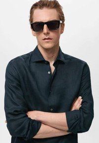 Gafas de sol negras con montura gruesa combinadas con una camisa de lino azul oscuro abotonada. El modelo cruza los brazos, mostrando una pose relajada.
