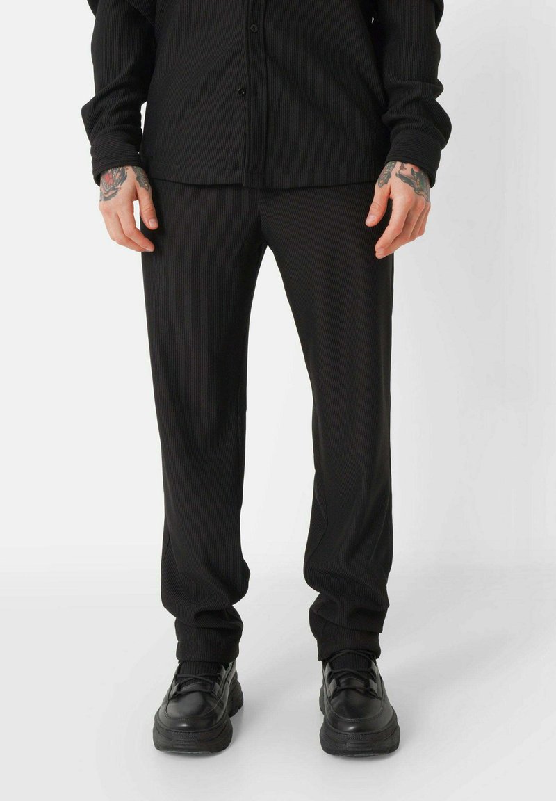 Zwarte geribde stoffen broek met een slim fit, met een gestructureerd oppervlak en elastische boorden. Gecombineerd met zwarte sportschoenen.