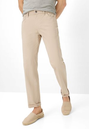 Hombre con pantalones beige remangados, zapatos tipo mocasín a juego y camiseta gris, de pie frente a un fondo blanco liso.