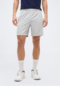 Άνδρας που φοράει ανοιχτό γκρι αθλητικά σορτς, λευκές ψηλές κάλτσες, άσπρα αθλητικά παπούτσια και σκούρο μπλε μπλουζάκι, όρθιος σε λευκό φόντο.