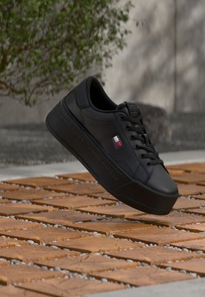 Czarny sneaker Tommy Jeans z naszywką z logo i tłoczonym brandingiem, unoszący się nad drewnianą i kamienną podłogą na zewnątrz, z rozmytym, zielonym tłem.