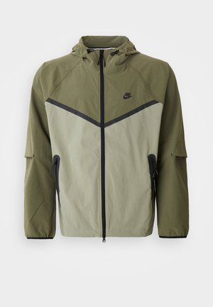 Herren Nike-Jacke in Olivgrün und Hellgrau, mit Reißverschluss, Kapuze und schwarzen Akzenten. Hergestellt aus leichtem, strukturiertem Stoff.