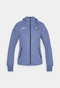 Blå zip-up jakke laget av mykt materiale, med hette, sidelommer og et logo på brystet. Inkluderer svart glidelås og detaljer.