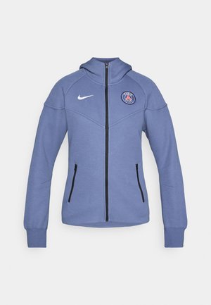 Blå zip-up jakke laget av mykt materiale, med hette, sidelommer og et logo på brystet. Inkluderer svart glidelås og detaljer.