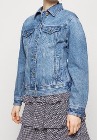 Veste en jean bleue au design classique, col pointu, poches avant, quincaillerie dorée et texture légèrement usée.