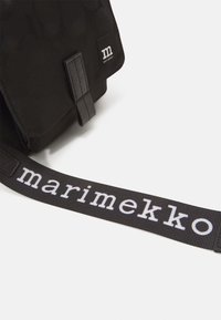 Black fabric crossbody bag with a textured pattern, featuring a branded strap in black and white with "marimekko" text and a flap closure.