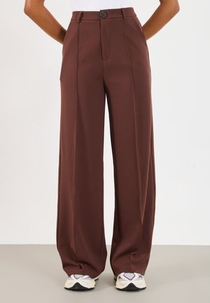 Personne portant un pantalon marron taille haute à jambes larges avec des coutures devant, associé à des baskets blanches et violettes, debout devant un fond uni.