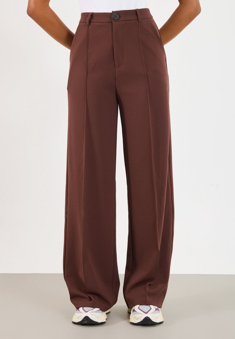 Personne portant un pantalon marron taille haute à jambes larges avec des coutures devant, associé à des baskets blanches et violettes, debout devant un fond uni.