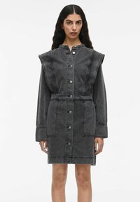 Femme portant une robe en denim noir délavé avec des manches longues, de grandes épaulettes, des boutons sur le devant, deux poches avant et une taille cintrée.