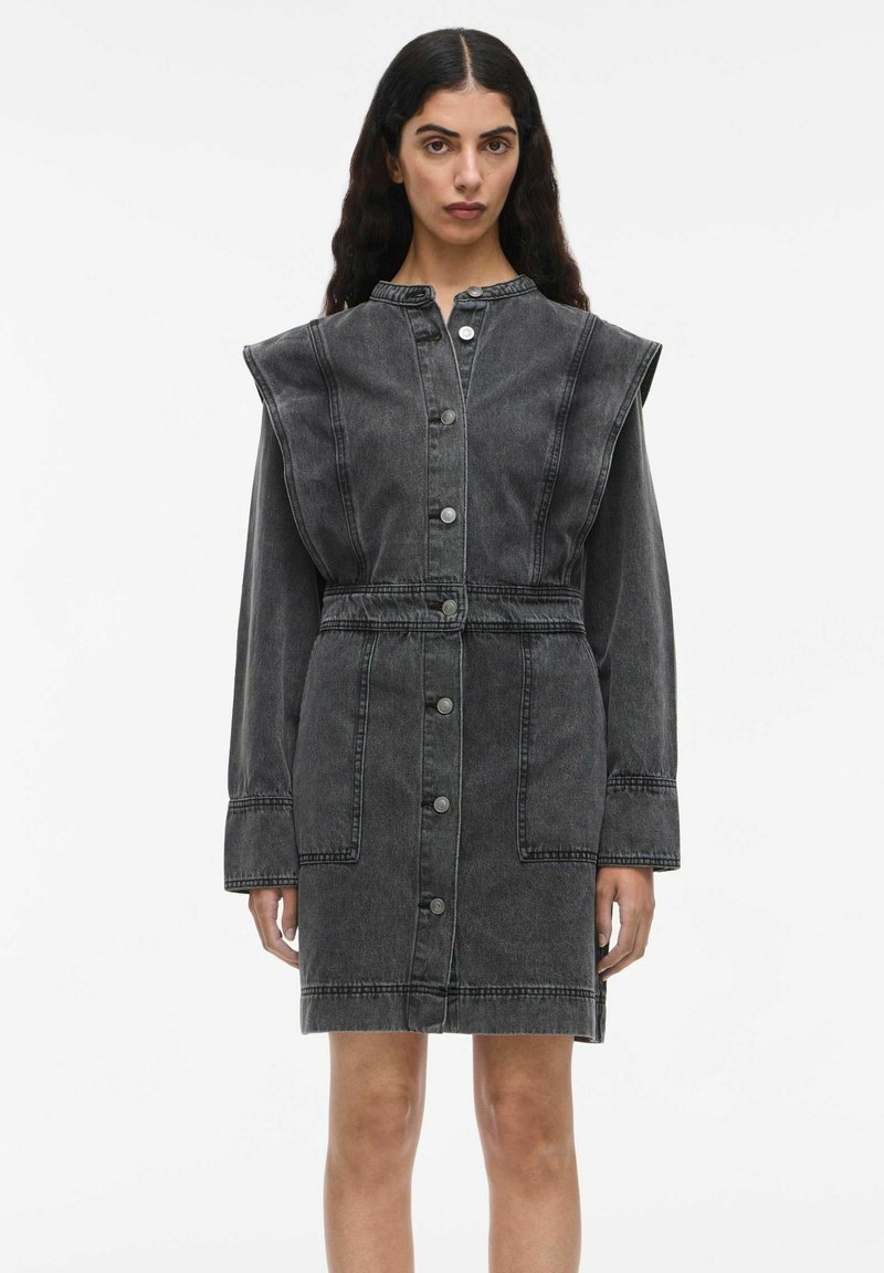 Femme portant une robe en denim noir délavé avec des manches longues, de grandes épaulettes, des boutons sur le devant, deux poches avant et une taille cintrée.