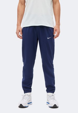 Personne portant un pantalon de survêtement Nike bleu marine, un t-shirt de sport en mesh blanc et des chaussures de sport blanches avec des semelles bleues et noires.