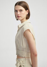 Beige cropped shirt met een kraag, korte mouwen en geplooide details bij de taille. Glad, licht glanzende textuur.