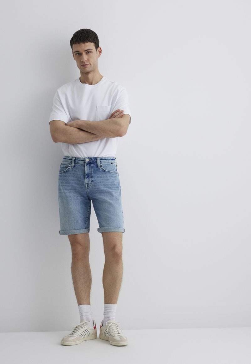Light blue denim shorts with rolled cuffs, paired with a plain white crewneck t-shirt. Shoes are beige with red accents and white socks.