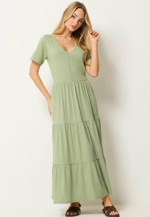 Donna con capelli biondi lunghi che indossa un maxi vestito verde chiaro a balze con scollo a V e maniche corte, posa su uno sfondo beige.
