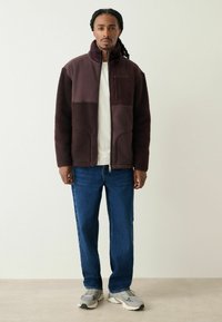 Fleece jacket in dark burgundy with a high collar, textured fabric, and a chest pocket. Paired with blue jeans and gray sneakers.