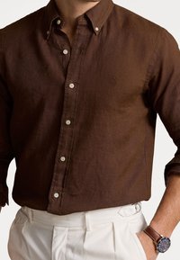 Camicia button-up in lino marrone con colletto e bottoni bianchi a contrasto. Presenta un piccolo logo ricamato sul petto. Abbinata a pantaloni chiari.