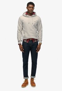 Grijze katoenen hoodie met trekkoorden over een rood geruit shirt, gedragen met donkere spijkerbroeken en bruine leren laarzen. Mouwen opgerold.