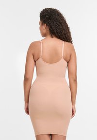 Frau mit lockigem Haar, von hinten gesehen, in einem beigen, figurbetonten Shapewear-Kleid mit dünnen verstellbaren Trägern vor einem weißen Hintergrund.
