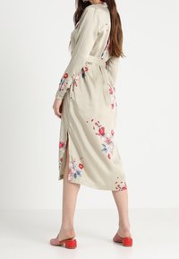 Lungo abito beige con motivi floreali in rosso e blu. Presenta una cintura, una spacco laterale e maniche lunghe. Indossato con sandali rossi con tacco a blocco.