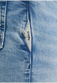 Närbild av ljusblå denimjeans söm med en vit tygetikett som står "CHASIN'" sydd i fickområdet.