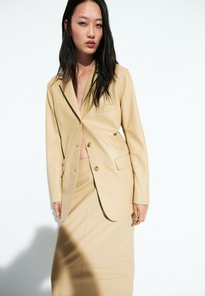 Frau mit langen dunklen Haaren, die eine beige maßgeschneiderte Jacke und einen passenden Rock trägt, steht vor einem weißen Hintergrund.