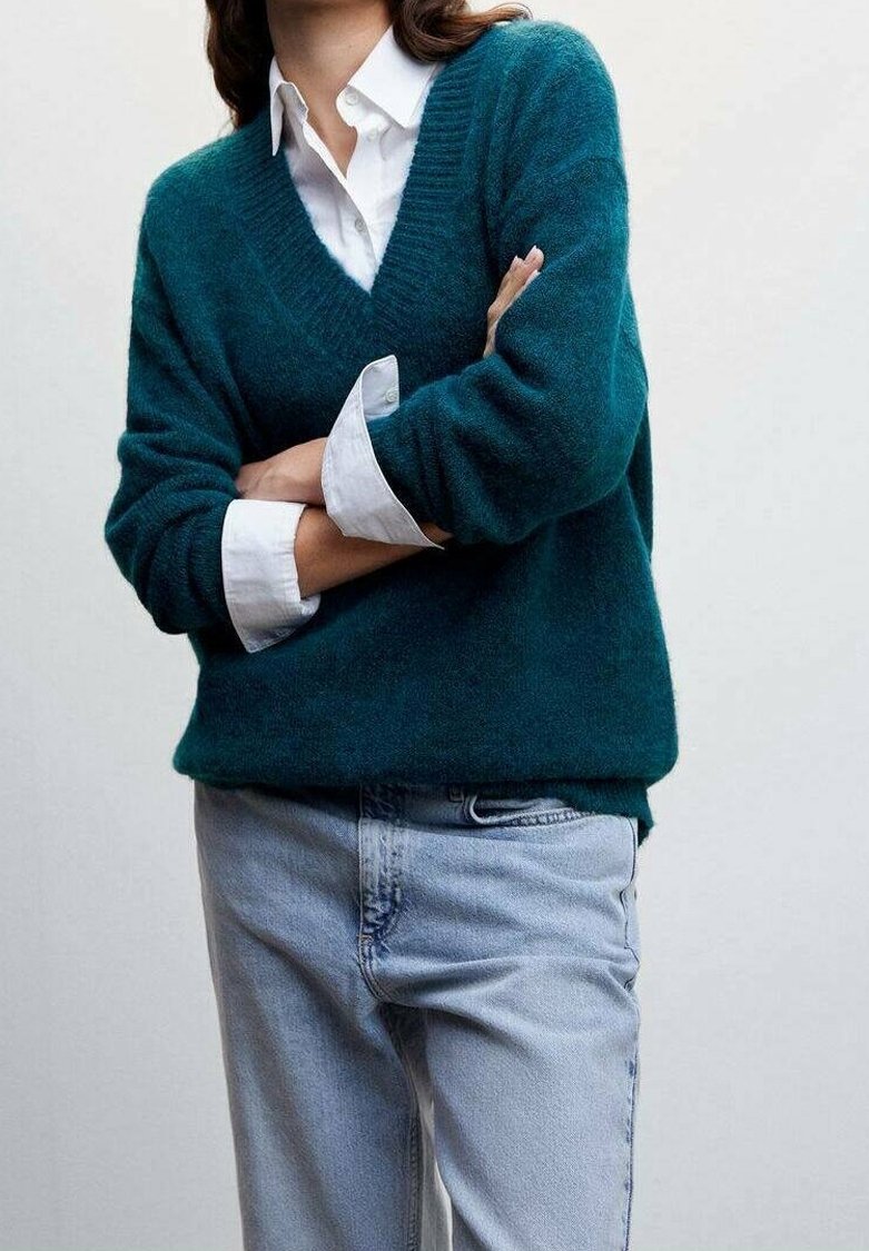 Personne portant un pull col en V couleur sarcelle sur une chemise blanche à boutons avec les manches retroussées, associée à un jean bleu clair, bras croisés.